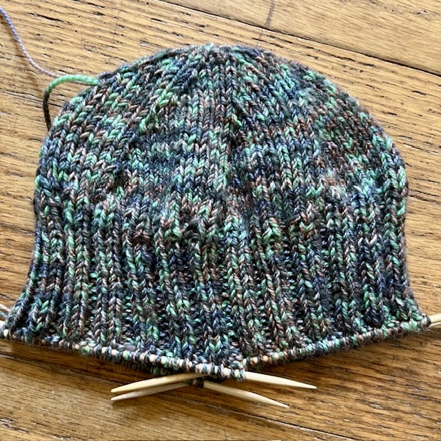 The top of the hat in smooth knitting, with a kind of textured spiral. The hat is mottled green, grey, and brown. The sides are knit in ridges. The hat is clearly not finished and there are still knitting needles in stitches at the bottom edge.