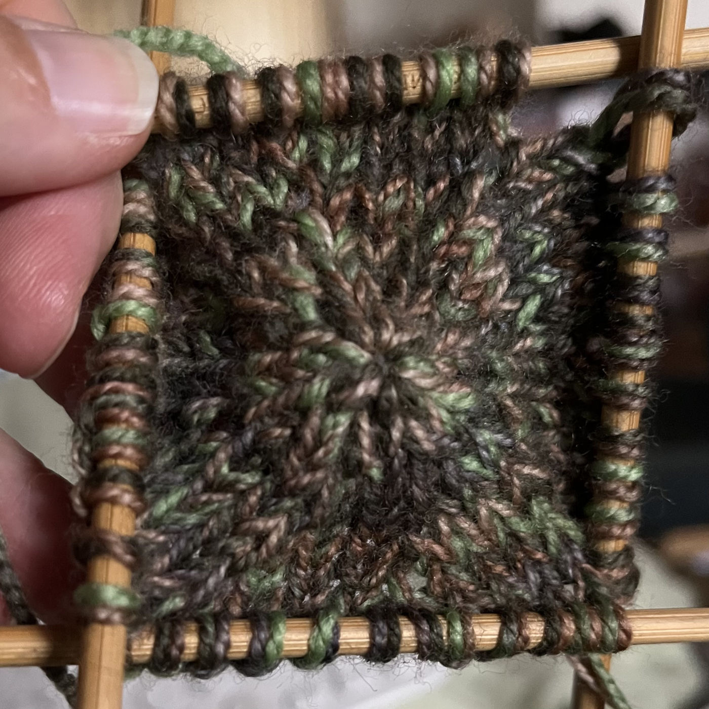 The center of the top of a hat on four bamboo knitting needles, being held up by a white person’s hand. Holding the two yarns together has jumbled the browns, grey, and greens together so that it looks a little like camouflage fabric.